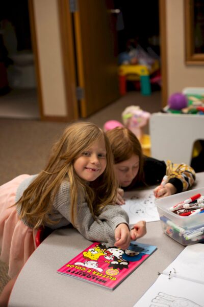 Two girls color at a table with crayons and activity books in a classroom setting.