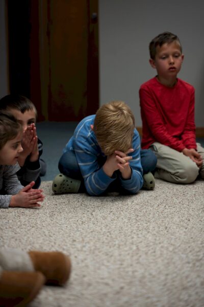 Four young boys sit on a carpet; one in the center is bowing his head with hands clasped.