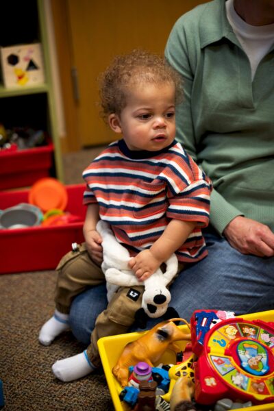 Toddler holding stuffed dog, sitting on adult's lap, surrounded by colorful toys.