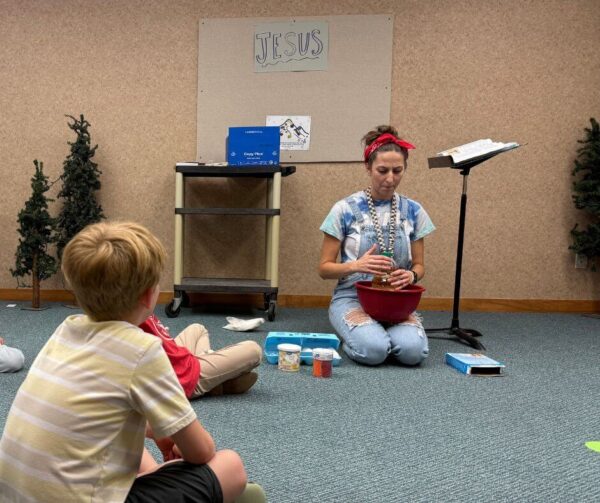 Woman kneeling, mixing ingredients in a bowl, teaches children in a classroom with "Jesus" sign.