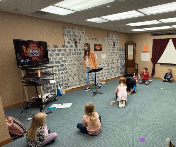A teacher talks to kids sitting on the carpet in a classroom with a TV and wall decorations.
